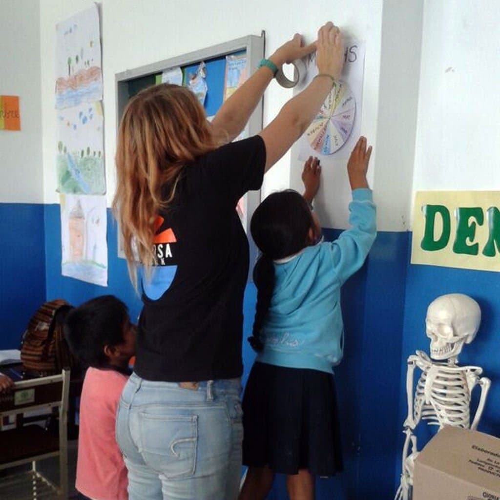 Ein:e Freiwillige:r hilft einem Kind bei einer Englischübung an der Wand des Klassenzimmers
